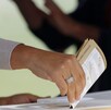 Imagen en primer plano de una mano colocando una boleta electoral dentro de una urna. El fondo es desenfocado.