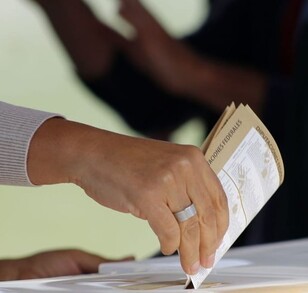 Imagen en primer plano de una mano colocando una boleta electoral dentro de una urna. El fondo es desenfocado.