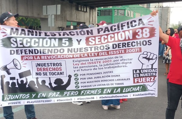 Se observa a personas de pie con una pancarta en las manos, visten playera roja.