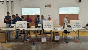 Se observa a varias personas de pie y tienen unas urnas de votar adelante.