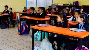 Se observa a varios alumnos en sus pupitres dentro de un salón de clases.