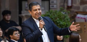 Exalcalde de Uruapan, Ignacio Campos sonriendo mientras sostiene un micrófono y durante una presentación pública con personas desenfocadas en el fondo