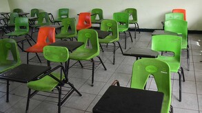 Mesabancos escolares con sillas color verde y naranjas dentro de un salón de clases vacío por suspensión de clases