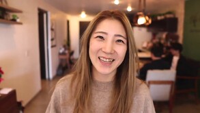 Youtuber Yuna Kim del canal Cocinando Corea sonriendo de frente a la cámara dentro de una cafetería con personas desenfocadas en el fondo