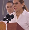 Mujer de lentes hablando ante una tribuna.