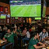 Se observa a varias personas sentadas dentro de un bar, tomando y comiendo; hay pantallas de televisión.