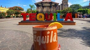 Cantarito con una rodaja de naranja delante de las letras turísticas de Tequila en medio de la plaza principal con un kiosco y la catedral