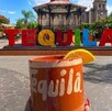 Cantarito con una rodaja de naranja delante de las letras turísticas de Tequila en medio de la plaza principal con un kiosco y la catedral