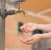 Llave de agua con un pequeño chorro saliendo y una persona extendiendo las manos debajo del líquido