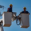 Dos trabajadores de la CFE en canastillas de una grúa realizando trabajos en una torres de luz y cables detrás de ellos
