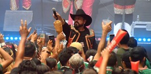 Cantante de banda extendiendo el micrófono hacia el público del Zona Fan durante el inicio del Mundial en Zapopan con músicos en el fondo