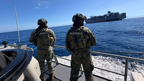 Dos militares de la Marina vistos de espaladas a bordo de un barco en altamar con un buque mercante con contenedores delante