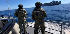 Dos militares de la Marina vistos de espaladas a bordo de un barco en altamar con un buque mercante con contenedores delante