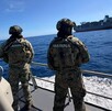 Dos militares de la Marina vistos de espaladas a bordo de un barco en altamar con un buque mercante con contenedores delante