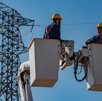 Dos trabajadores de la CFE en canastillas de una grúa realizando trabajos en una torres de luz y cables detrás de ellos