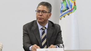 Hombre de traje y lentes sentado junto a una bandera de Puebla.