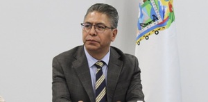Hombre de traje y lentes sentado junto a una bandera de Puebla.