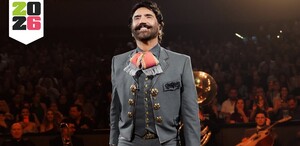 Cantante Alejandro Fernández con traje de charro sonriendo durante un concierto en un palenque con el público y músicos en el fondo