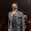 Cantante Alejandro Fernández con traje de charro sonriendo durante un concierto en un palenque con el público y músicos en el fondo