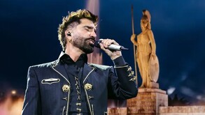 Alejandro Fernández cantando con un micrófono en la mano y una imagen de La Minerva desenfocada en el fondo