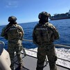 Dos militares de la Marina vistos de espaladas a bordo de un barco en altamar con un buque mercante con contenedores delante