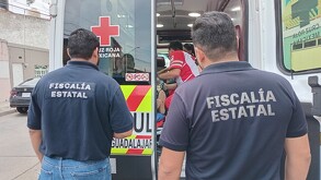 Agentes de la Fiscalía de Jalisco vistos de espaldas con una ambulancia de la Cruz Roja con paramédicos dando atención a una mujer de la tercera edad