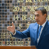 El político Zamora habla en un podio con un traje azul y corbata. Detrás de él, hay una pared conmemorativa con fechas históricas y una bandera del Estado