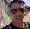 Hombre de gafas y playera negra hablando a una cámara en un día soleado.