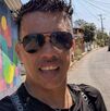 Hombre de gafas y playera negra hablando a una cámara en un día soleado.