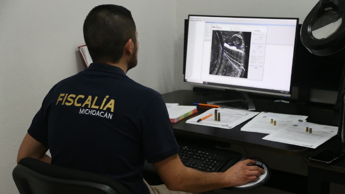 Policía investigador de la Fiscalía de Michoacán visto de espaldas mientras trabaja en una computadora con documentos delante