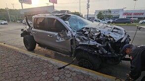 Camioneta RAM de lujo destruida de la parta frontal y trasera siendo remolcada por una grúa con un policía vial delante y una plaza comercial en el fondo