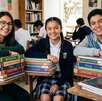 Tres estudiantes de diferentes edades sentados frente a una mesa con varias pilas de libros en un salón de clases