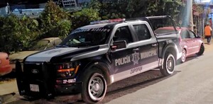 Vista lateral de noche de una patrulla de la Policía de Zapopan frente a unas plantas y con automóviles alrededor