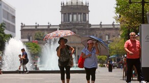 Mujeres mayores caminando son sombrillas para protegerse del sol en el centro de Guadalajara con el Hospicio Cabañas desenfocado en el fondo