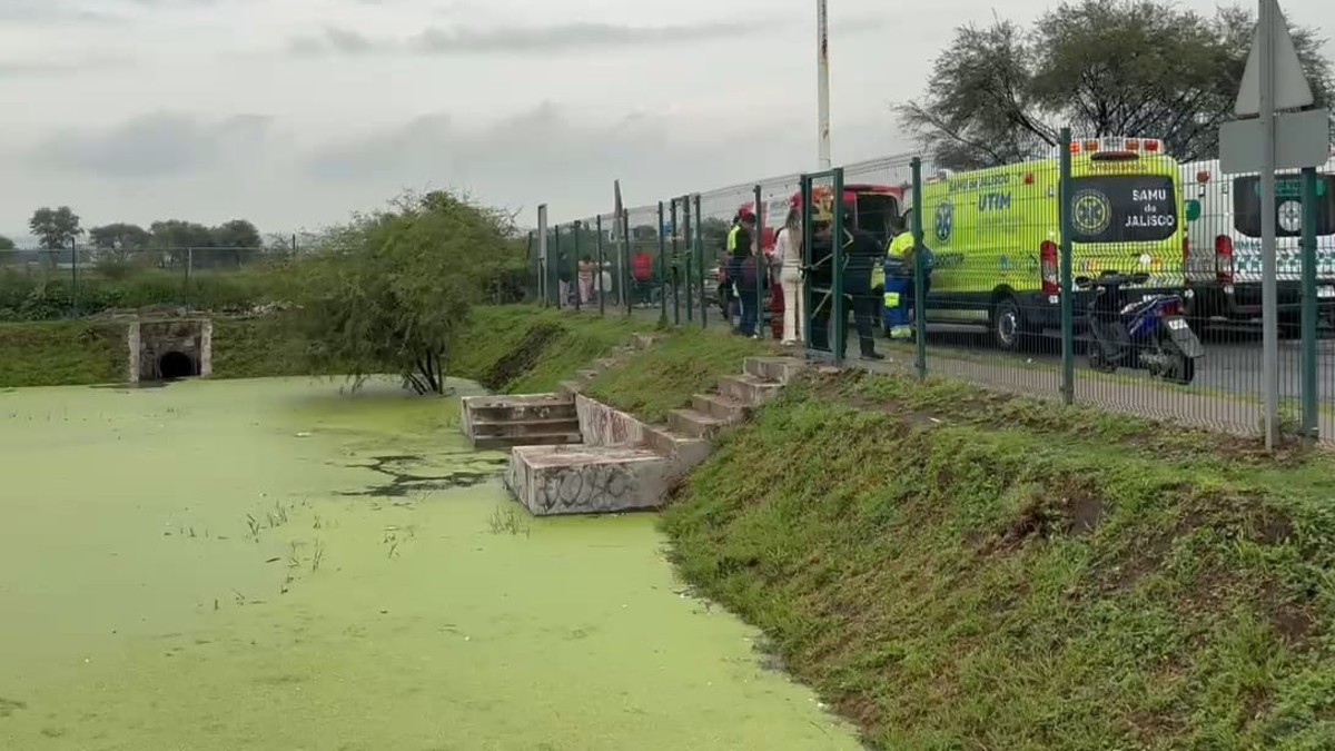 Vaso regulador con agua estancada de color verde dentro de una cancha hundida con paramédicos, policías y ambulancias al lado del enrejado
