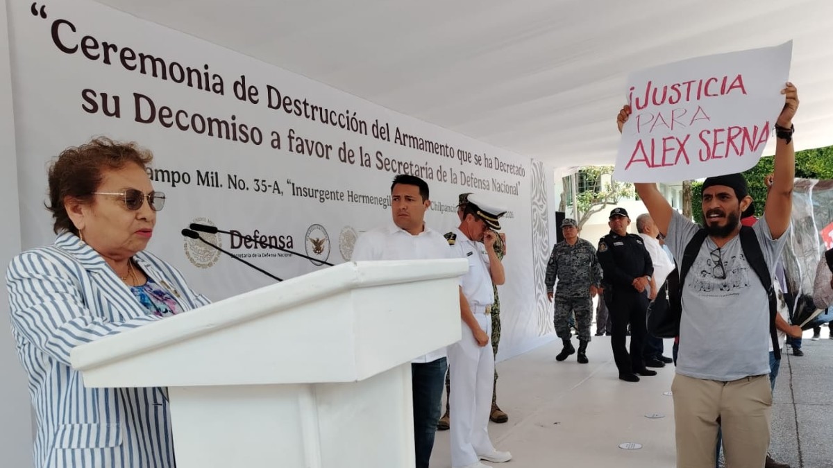 Hombre protestando en un evento oficial, mientras una mujer habla frente al micrófono.