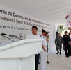 Hombre protestando en un evento oficial, mientras una mujer habla frente al micrófono.