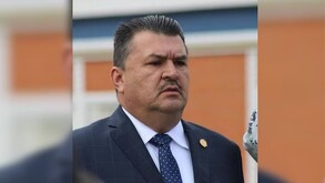 Julio Meza Gaona visto de perfil con el seño fruncido mientras viste de traje delante de un fondo naranaja