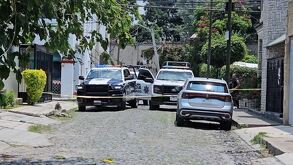 Patrullas de la Policía Municipal de Tlajomulco dentro de un área del crimen acordonada en una calle empedrara con autos y casa alrededor