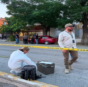 Se observa a un hombre y una mujer en la calle, una mujer vestida con traje blanco de perito y una cinta amarilla de precaución.