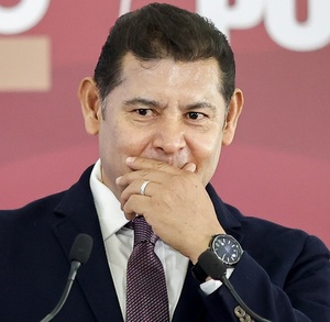 Hombre con traje se cubre la boca con las manos mientras habla ante un micrófono.