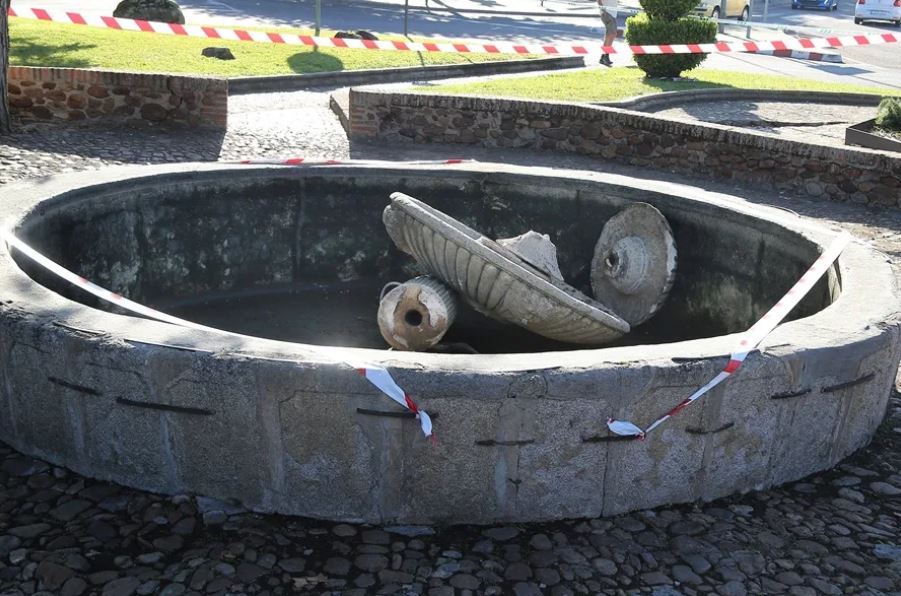 Fuente rota y acordonada por una cinta policiaca en medio de un suelo empedrado con plantas alrededor