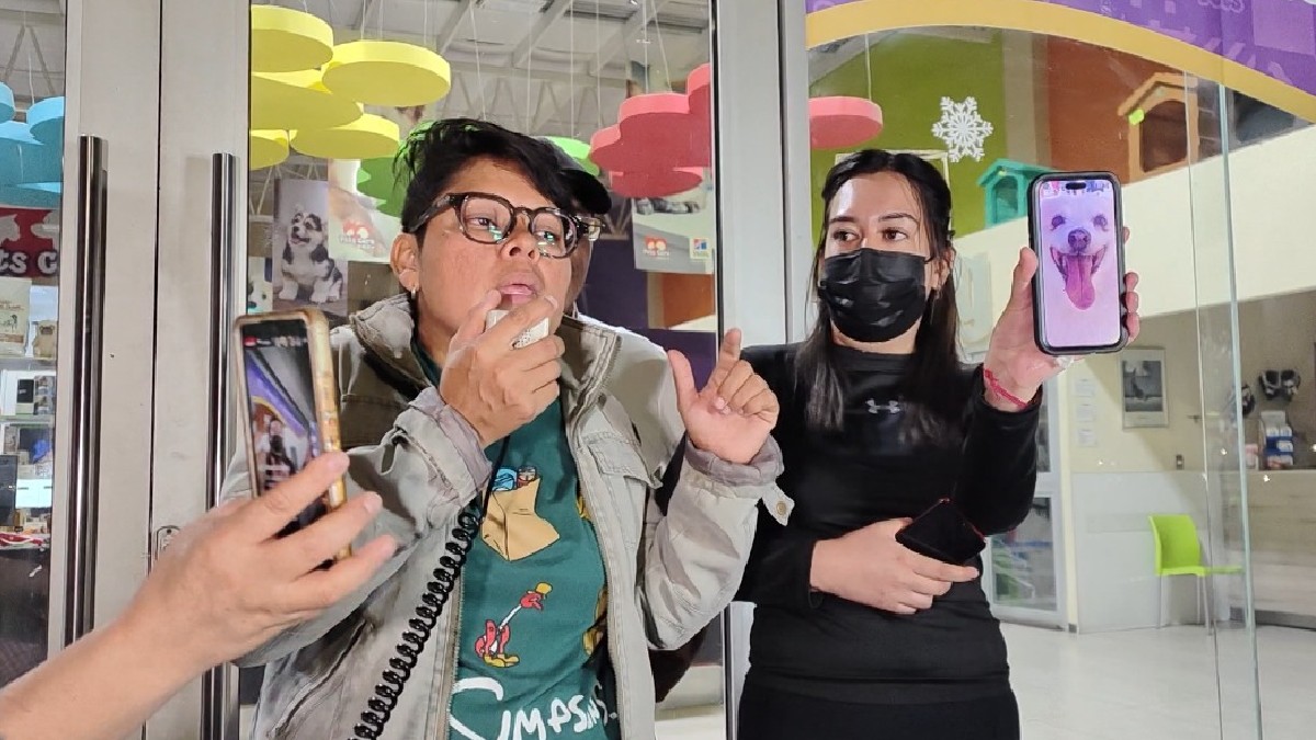 Se observa a dos mujeres, una con lentes y otra cargando un celular en la mano donde se ve un perrito color blanco.