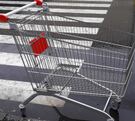 Carrito metálico de supermercado en medio de un estacionamiento con el suelo mojado por lluvia, un cruce peatonal y un poste