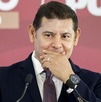 Hombre con traje se cubre la boca con las manos mientras habla ante un micrófono.