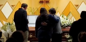 Personas abrazadas frente al ataúd de una persona muerta.