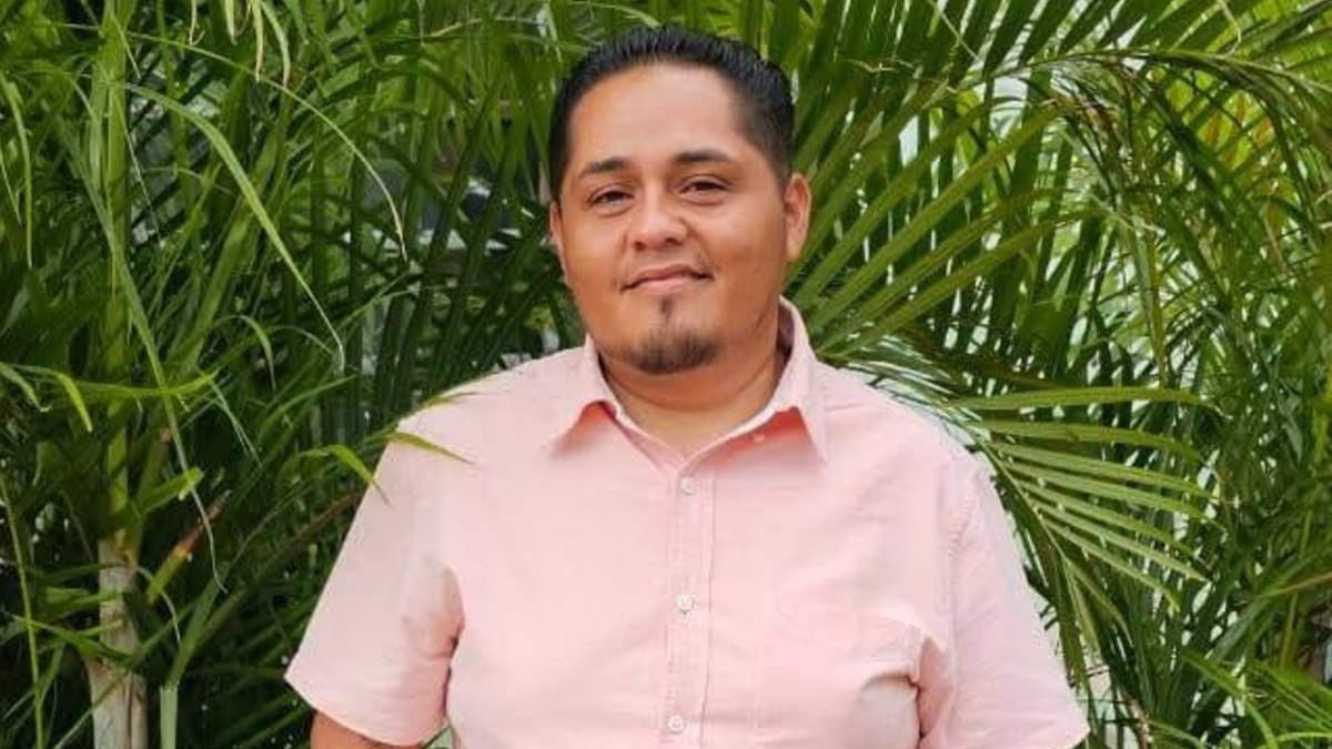 Hombre de camisa rosa posa para una fotografía junto a plantas largas.