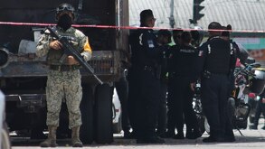 Militar armado parado junto a una cinta roja con policías.